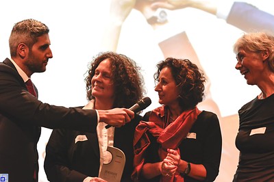 Ein junger Mann im Anzug ehrt drei fröhliche Frauen mit einem Mikrofon zum DEA Award