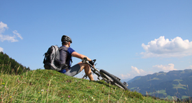 Ein junger Mann mit Fahrrad macht eine Pause auf einem grasigen Hügel mit Blick ins Tal