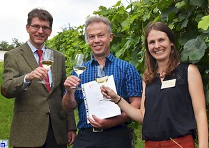 Dr. Martin Barth, der Preisträger Daniel Feuerstein und Andrea Gierden stehen vor Weinreben und stoßen mit dem ausgezeichneten Sekt an.