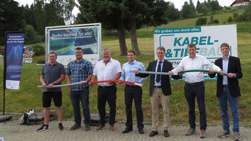 Die 7 Beteiligten stehen vor Werbetafeln und halten Breitbandkabel.